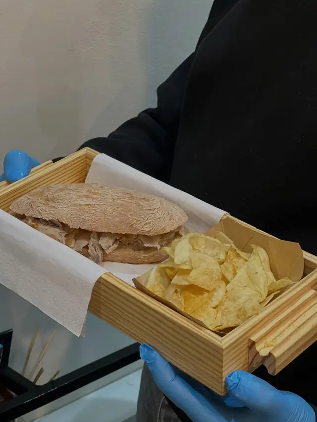Caixa de madeira com sandes de leitão e batatas fritas, segura por mãos de luvas